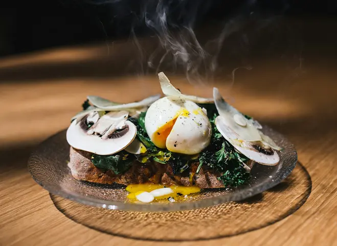 A slice of toasted bread topped with sautéed greens, raw mushroom slices, and a poached egg with runny yolk, served on a glass plate with steam rising.